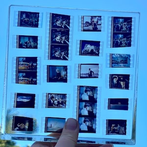 May include: A clear, rectangular film strip holder containing multiple black and white film frames. The frames depict scenes from a movie, including people, cars, and landscapes. The holder is held against a blue sky.