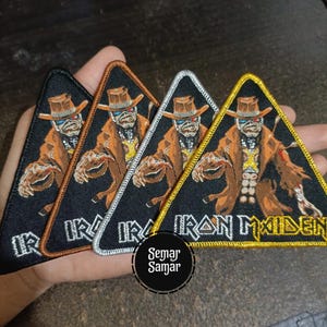 Puede incluir: Cuatro parches triangulares de Iron Maiden con Eddie, la mascota, con abrigo y sombrero marrón. Los bordes son negros, marrones, plateados y dorados. El texto "IRON MAIDEN" está en la parte inferior.