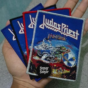 Op de afbeelding: Verschillende vierkante patches met de bandnaam "Judas Priest" in het wit, en de albumtitel "Painkiller". De patches tonen een afbeelding van een gevleugeld wezen op een motorfiets. Ze hebben blauwe achtergronden met rode, zwarte en witte randen.