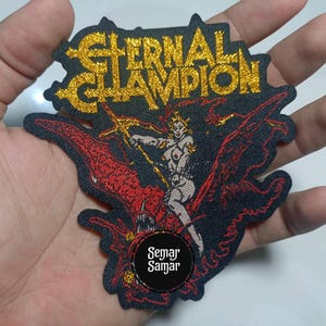 May include: Embroidered patch with the words "Eternal Champion" in gold glitter lettering. The patch features a warrior woman riding a red dragon, with the text "Semar Samar" in a circle at the bottom. The patch is black with red and gold accents.