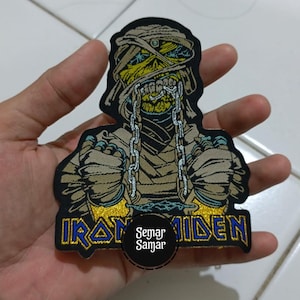 May include: Embroidered patch depicting a mummy-like figure with a yellow face and bandages, wearing a chain. The patch features the words "IRON MAIDEN" in blue and gold. A circular logo with the text "Semar Samar" is also present.