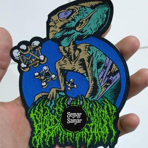 Può includere: Una toppa colorata con un alieno con la pelle verde, gli occhi viola e il corpo marrone. L'alieno è ambientato su uno sfondo blu con strutture molecolari bianche e nere. La toppa ha il testo "Semar Samar".