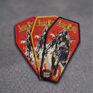 Puede incluir: Tres parches bordados en forma de ataúd con el texto Judas Priest en letras doradas. Cada parche muestra el gráfico de una figura con armadura, escudo y espada sobre un fondo rojo, con bordes distintivos en colores rojo, amarillo y negro.