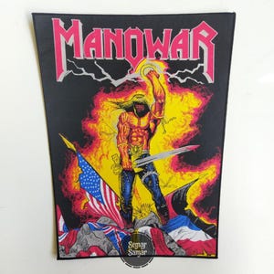 May include: A rectangular fabric patch featuring the word MANOWAR in large pink letters at the top. Below, a muscular figure with long hair holds a sword aloft against a yellow and orange flame background, with various national flags below.