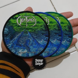 May include: Three circular embroidered patches featuring the band name "Obituary" in silver lettering. The patches depict a blue and green landscape with the text "frozen in time". Each patch has a different coloured border: black, blue, and green.