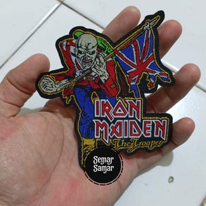 Op de afbeelding: Geborduurde patch met de Iron Maiden-mascotte, Eddie, in een rode jas en blauwe broek, met een Union Jack-vlag. De patch bevat de bandnaam en de tekst "The Trooper" in een gestileerd lettertype, met een zwarte rand.