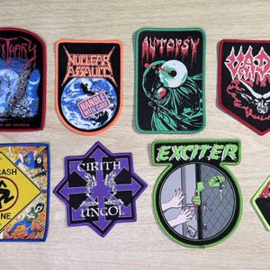 May include: A collection of eight embroidered fabric patches featuring various band logos and graphic designs, including Obituary, Nuclear Assault, Autopsy, Vader, D.R.I. Thrash Zone, Cirith Ungol, Exciter, and Overkill, arranged in two rows.