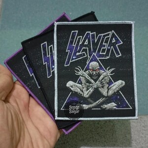 May include: A rectangular black fabric patch featuring the white text SLAYER above a graphic of a pale, red-eyed figure with crossed arms inside a triangle. The background is a dark purple and black starry space design with a white stitched border.