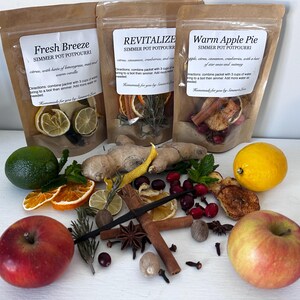 May include: Three brown paper pouches labeled "Fresh Breeze", "Revitalize", and "Warm Apple Pie" simmer potpourri. Ingredients like citrus, cinnamon, cranberries, and apples are arranged around the pouches. Fresh fruit and spices are also visible.
