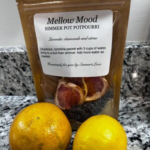 May include: A clear bag of "Mellow Mood" simmer potpourri with dried citrus, lavender, and chamomile. Two whole citrus fruits, a lemon and a tangerine, sit in front of the bag. The bag's label includes instructions and the phrase "Homemade for you by Simmer & Love."