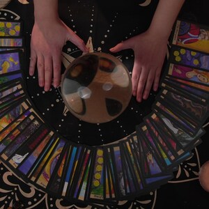 May include: A crystal ball sits on a black tablecloth surrounded by a spread of tarot cards. Two hands are placed on either side of the ball. There are candles and other decorative items on the table.