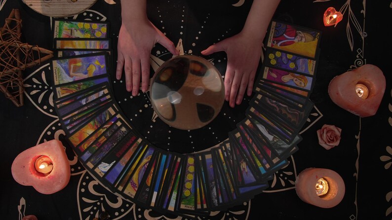 May include: A crystal ball sits on a black tablecloth surrounded by a spread of tarot cards. Two hands are placed on either side of the ball. There are candles and other decorative items on the table.