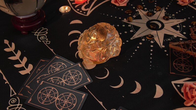 May include: A black cloth with a gold star and moon design. There are tarot cards with gold accents, a clear crystal, and a small candle on the cloth.