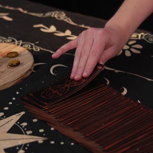 May include: A person's hand shuffles a deck of tarot cards on a black and gold patterned table cloth. The cards have a dark wood finish with gold accents.