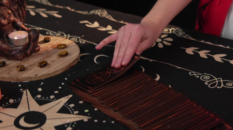 May include: A person's hand shuffles a deck of tarot cards on a black and gold patterned table cloth. The cards have a dark wood finish with gold accents.