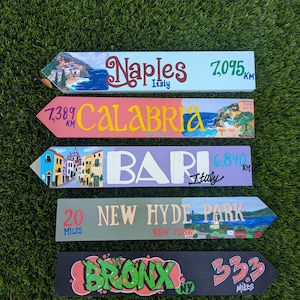 May include: Five wooden directional signs featuring painted illustrations and text. The signs read: Naples Italy 7,095 KM, 7,389 KM Calabria Italy, Bari Italy 6,840 KM, 20 Miles New Hyde Park New York, and The Bronx NY 33.3 Miles with graffiti-style lettering.