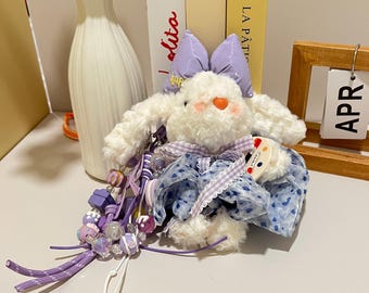 May include: A plush white bunny keychain with a lavender bow and blue patterned dress. The bunny has a pink nose and a beaded charm with purple and clear beads. A small wooden calendar displays "APR".