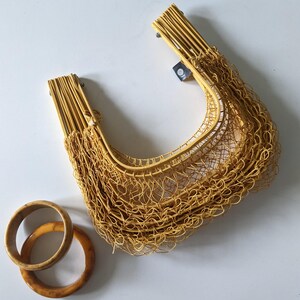 May include: A woven yellow handbag with a net-like design, featuring two wooden handles. Two wooden bangles in shades of brown and tan are placed near the bag. The bag is made of thin, flexible material.