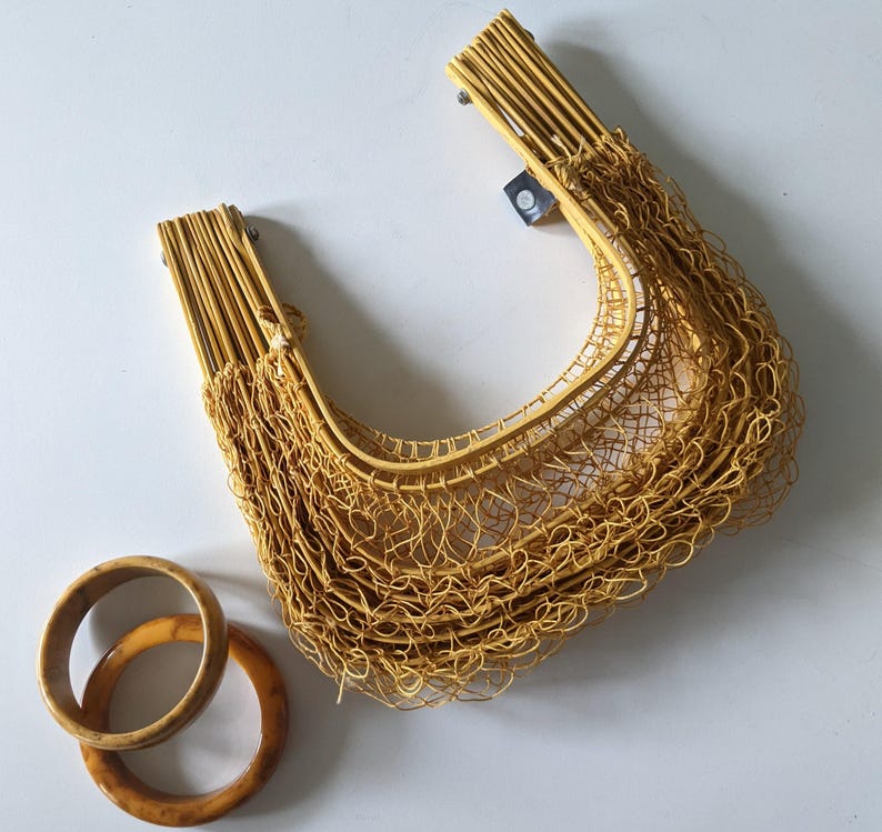 May include: A woven yellow handbag with a net-like design, featuring two wooden handles. Two wooden bangles in shades of brown and tan are placed near the bag. The bag is made of thin, flexible material.
