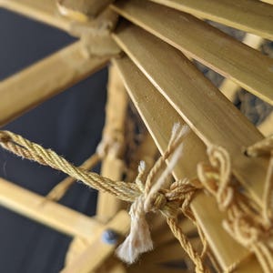 May include: Close-up of a bamboo structure with a woven design. The structure is held together with a twisted, light-brown rope. The bamboo is a light tan color, and the background is dark.