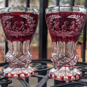 May include: Two matching ruby red and clear cut glass goblets featuring an intricate etched floral pattern surrounding the engraved initials H.E.K. The stems and bases display a clear glass diamond-cut lattice design with a scalloped edge.