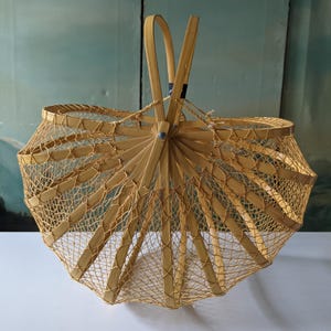 May include: A light brown, open-weave basket with a handle. The basket has a unique, umbrella-like shape, with a series of wooden ribs radiating from a central point. The sides are constructed with a fine, woven mesh, creating an airy and decorative design.