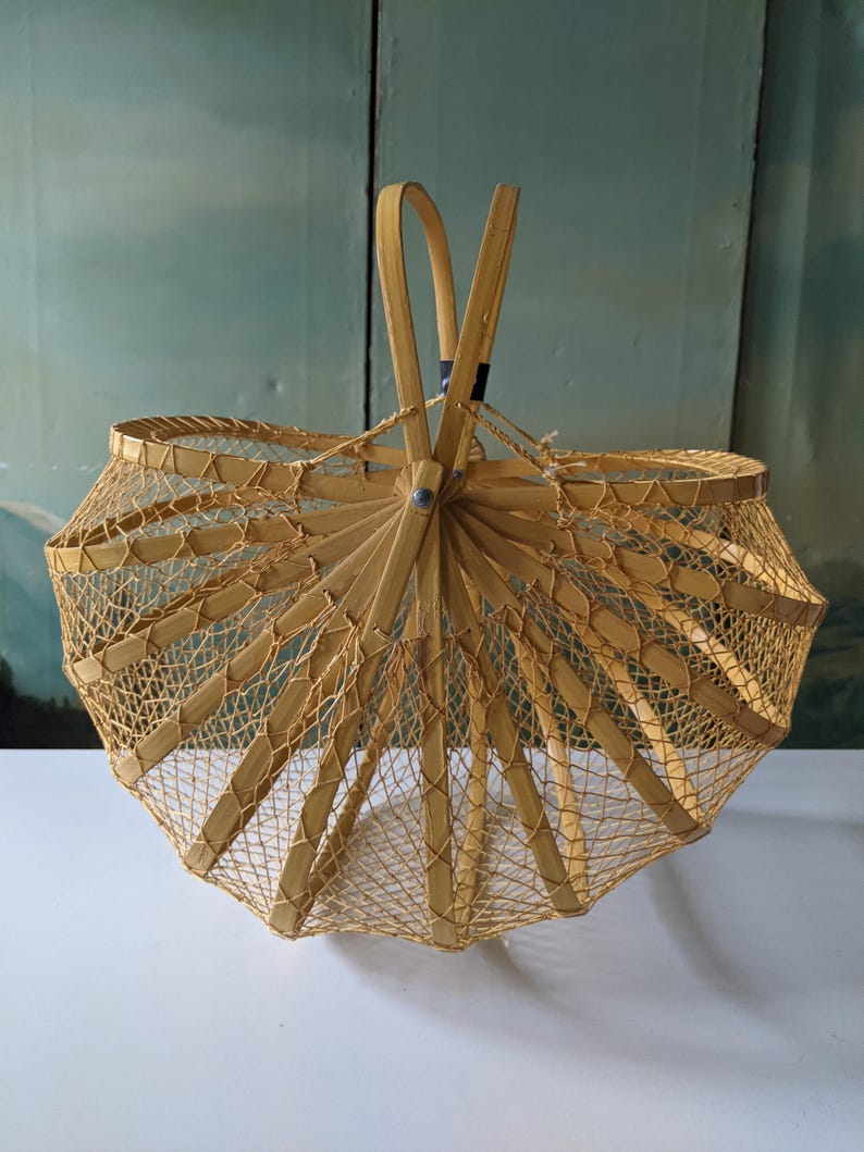 May include: A light brown, open-weave basket with a handle. The basket has a unique, umbrella-like shape, with a series of wooden ribs radiating from a central point. The sides are constructed with a fine, woven mesh, creating an airy and decorative design.