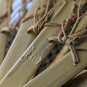 May include: Close-up of several bamboo fan blades, painted a light yellow color. The blades are connected with tan-colored string, knotted at the top. The image has a shallow depth of field, with the focus on the fan blades.