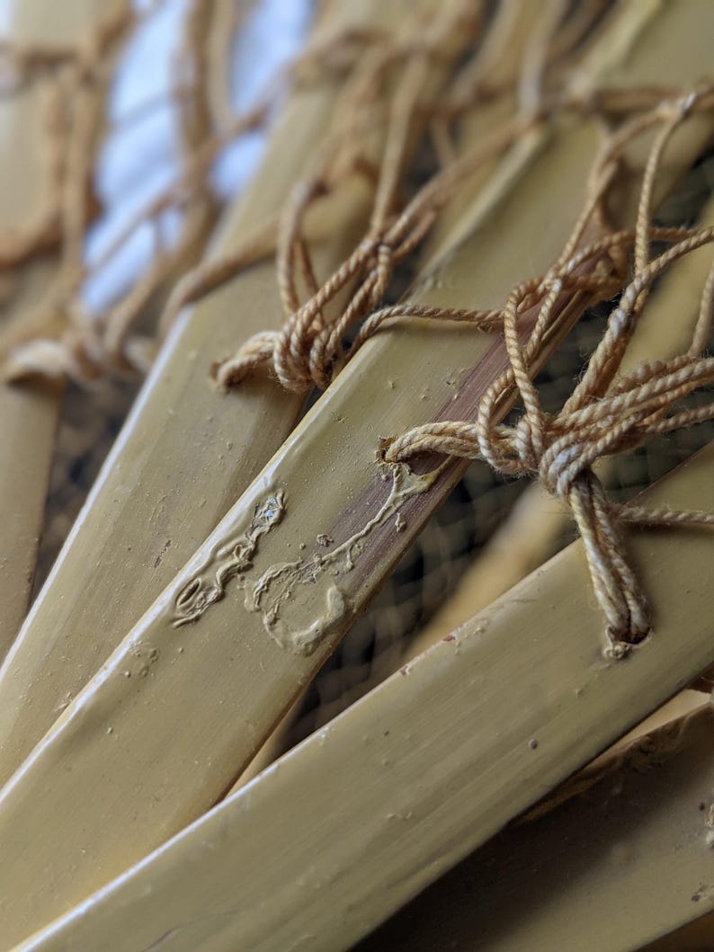 May include: Close-up of several bamboo fan blades, painted a light yellow color. The blades are connected with tan-colored string, knotted at the top. The image has a shallow depth of field, with the focus on the fan blades.