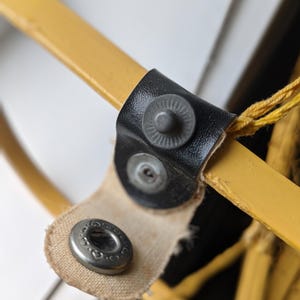 May include: Close-up of a yellow metal object with a black leather strap and metal snaps. The strap is attached to a yellow metal bar. A beige fabric is also attached to the metal snaps.