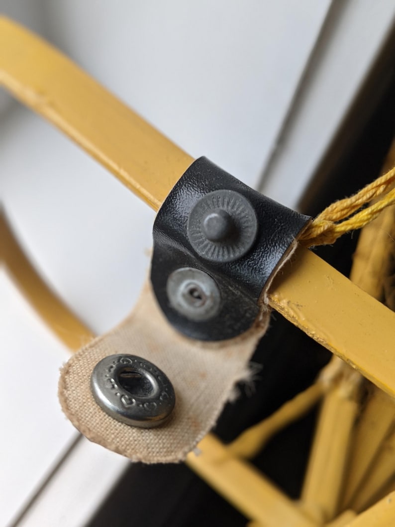 May include: Close-up of a yellow metal object with a black leather strap and metal snaps. The strap is attached to a yellow metal bar. A beige fabric is also attached to the metal snaps.