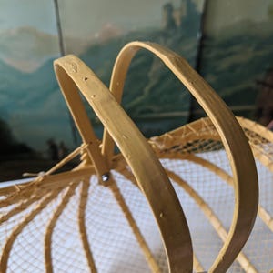May include: A close-up of a vintage, light-colored basket with a curved handle. The basket has a woven net design and a leather strap. The handle is made of a light-colored wood.