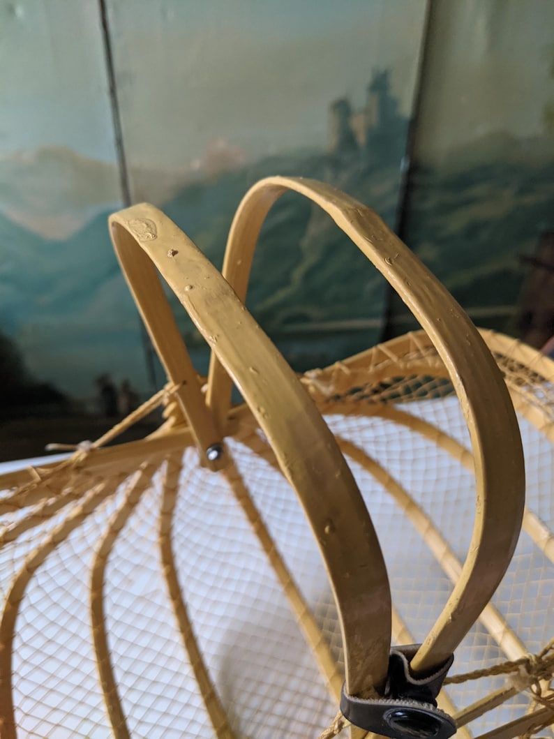 May include: A close-up of a vintage, light-colored basket with a curved handle. The basket has a woven net design and a leather strap. The handle is made of a light-colored wood.