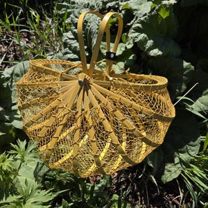 May include: A yellow, woven basket with a handle, set against a backdrop of green foliage. The basket's design features a lattice-like pattern, with a sturdy handle for carrying. The basket is likely used for gathering items.
