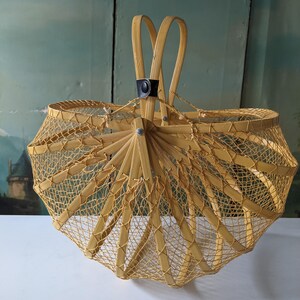 May include: A vintage, light yellow woven basket with a unique umbrella-like design. The basket features a sturdy handle and a mesh-like pattern throughout the body. The basket is made of bamboo and has a black leather accent.