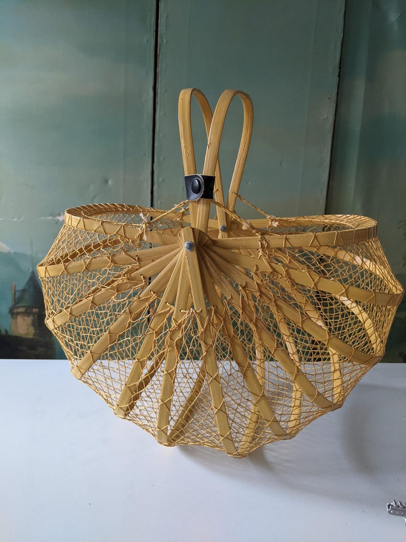 May include: A vintage, light yellow woven basket with a unique umbrella-like design. The basket features a sturdy handle and a mesh-like pattern throughout the body. The basket is made of bamboo and has a black leather accent.