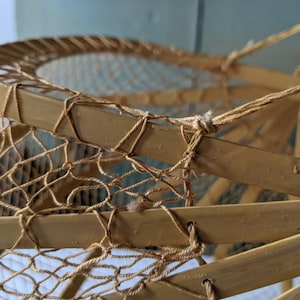 May include: Close-up of a vintage, handcrafted snowshoe. The frame is made of light brown wood, with a woven net of natural-colored twine. The twine is knotted to the frame, creating a sturdy and functional design. The snowshoe is a classic example of outdoor gear.