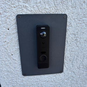 May include: A black rectangular video doorbell mounted on a dark gray metal plate with four corner screws. The device features a circular camera lens at the top, a small indicator light, a central button, and a speaker grille at the bottom.