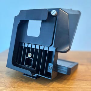 Könnte beinhalten: Schwarzer Kameramonitor mit einem klappbaren Sonnenschutz und einer Metallschraube in der oberen rechten Ecke. Der Monitor hat einen schwarzen Kunststoffrahmen und ein Gittermuster auf der Rückseite.