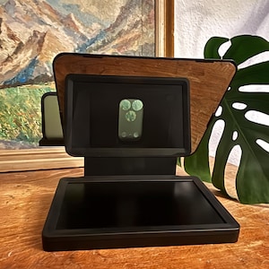 Peut inclure: Un support de téléphone noir et boisé avec une base rectangulaire et une zone d'affichage inclinée. Le support a un dos en bois et un cadre noir. L'écran du téléphone affiche une image verte. Le support est sur une surface en bois.