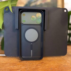 Könnte beinhalten: Eine schwarze Handyhülle mit durchsichtigem Rücken und einem kreisförmigen magnetischen Ladebereich wird in einem dunkelgrauen Halter angezeigt. Die Handyhülle ist auf einer dunkelgrauen rechteckigen Basis mit einem Loch in der Ecke montiert.