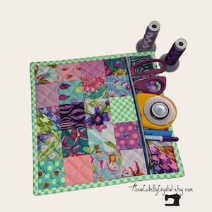 May include: A colorful patchwork sewing organizer with a zippered pocket, holding scissors, a rotary cutter, pens, and spools of thread. The organizer features a variety of floral and geometric patterns in shades of pink, teal, and purple, with a green checkered border.