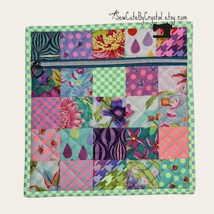 May include: A quilted square pouch with a zipper closure, featuring a patchwork design of various floral and geometric patterns in shades of pink, purple, green, and blue. The pouch has a green checkered border and the text "SewCuteByCrystal.etsy.com".