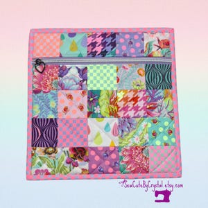 May include: A quilted square pouch with a zipper closure. The pouch is made of various patterned fabric squares in shades of pink, purple, green, and blue. The zipper pull is heart-shaped. The edges are trimmed with a pink and white checkered pattern.