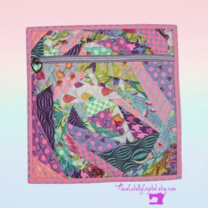May include: A quilted square pouch with a zipper closure. The pouch features a patchwork design with various fabrics in shades of purple, pink, green, and blue. The zipper pull is heart-shaped, and the edges are trimmed with a bright pink border.