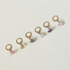 Puede incluir: Cinco pendientes de aro dorados, cada uno con un colgante de piedra preciosa de diferente color. Las piedras preciosas son rosa, amarillo-verdoso, azul-verdoso, morado y rosa. Los pendientes se muestran sobre una superficie blanca.