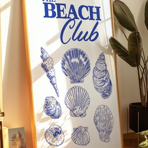 Puede incluir: Un cuadro enmarcado con borde dorado que presenta el texto "THE BEACH Club" en azul. La impresión muestra varias ilustraciones de conchas marinas azules sobre un fondo blanco, creando un tema costero.