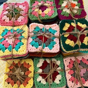May include: A stack of colorful, square crochet coasters tied with twine. Each coaster features a granny square design with various colors, including pink, green, yellow, and blue. The coasters are handmade and suitable for home decor.