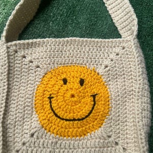 May include: A beige crocheted tote bag with a yellow smiley face design. The bag has a long strap and is made with a simple stitch pattern.