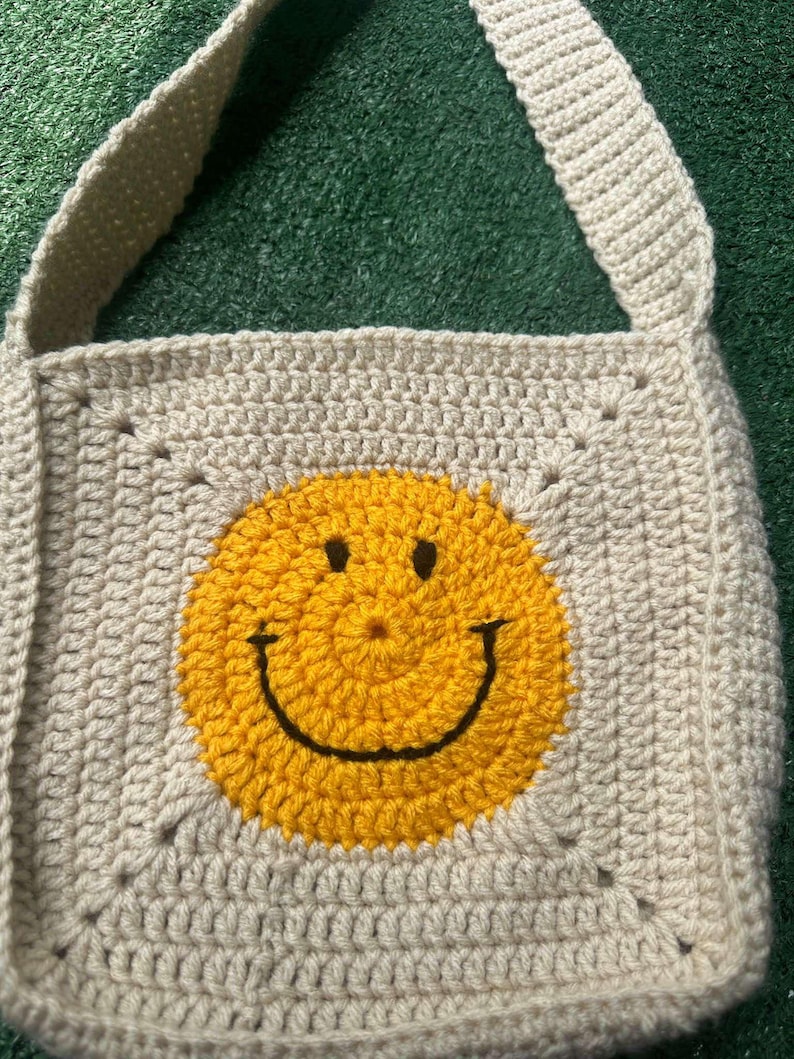 May include: A beige crocheted tote bag with a yellow smiley face design. The bag has a long strap and is made with a simple stitch pattern.